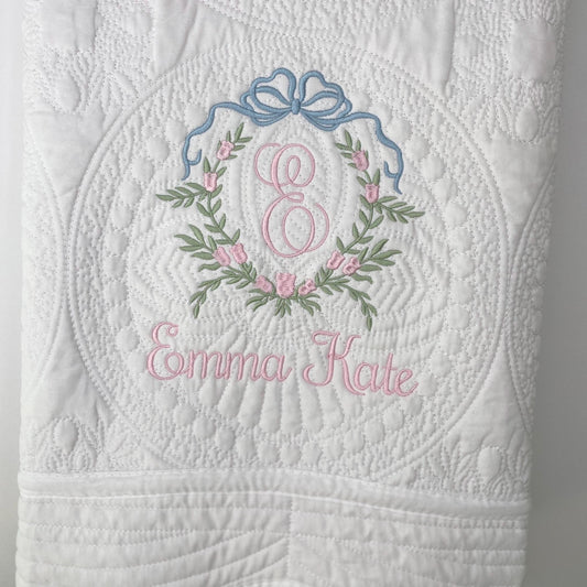 White quilt with detailed floral and bow embroidery in pink and blue, featuring a pink monogram "E" and the name Emma Kate, showcasing the Personalized Bow & Pink Floral Wreath Monogram Baby Quilt | Custom Embroidered Heirloom Blanket design.