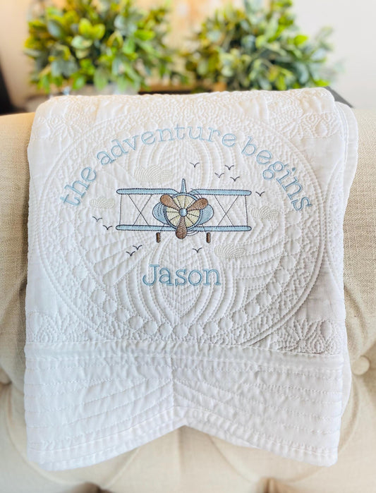 Personalized Vintage Airplane Quilt | Custom Name Heirloom Blanket featuring embroidered vintage airplane with the phrase "the adventure begins" and custom name Jason on white quilted fabric.