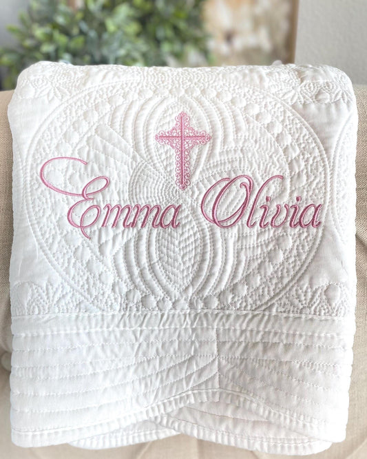 Folded white quilt with intricate embroidered patterns and a pink cross above the personalized names Emma Olivia, showcasing the Personalized Embroidered Cross Baptism Baby Quilt | Custom Name Christening Heirloom Keepsake.