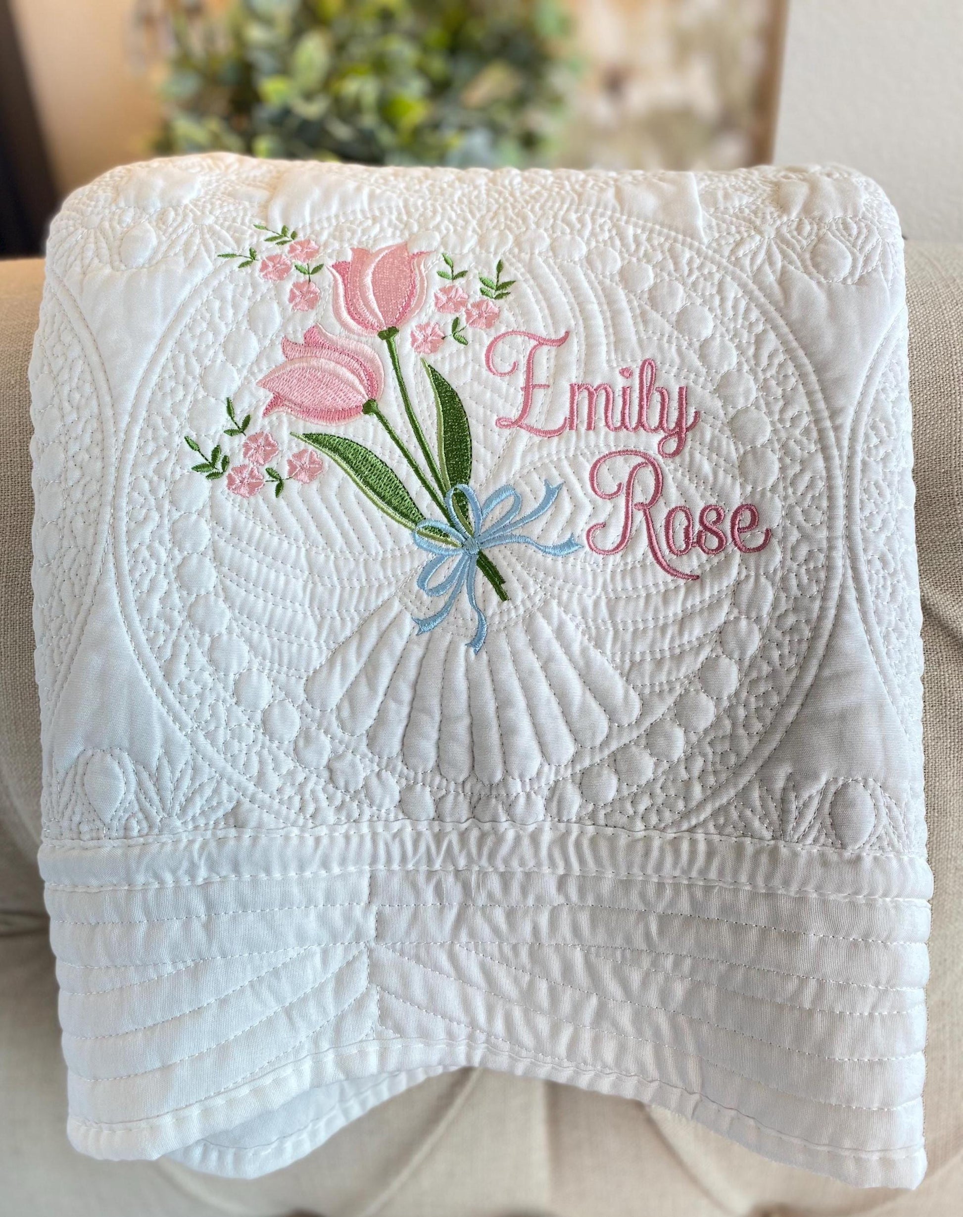 White quilt with intricate quilting featuring a personalized tulip bouquet and bow embroidery with the name Emily Rose in pink script on a soft background, showcasing Personalized Tulip Bouquet & Bow Baby Quilt | Custom Embroidered Heirloom Blanket for Girls.
