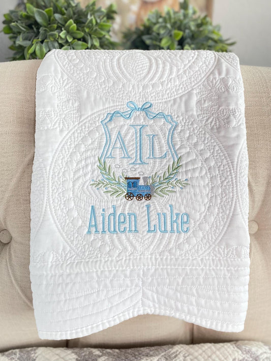 White quilt with intricate stitching, featuring a blue embroidered monogram, a small steam train, and the name Aiden Luke, showcasing the Personalized Monogram Steam Train Baby Quilt | Custom Name Heirloom Keepsake.