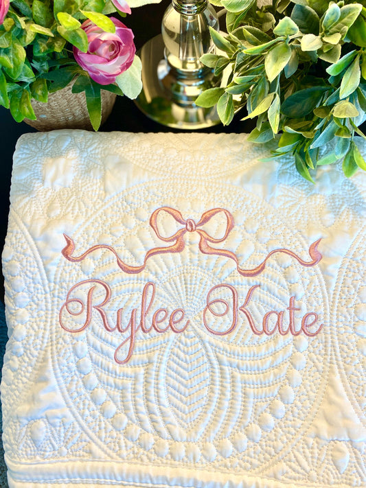 Personalized Pastel Pink Ribbon Bow Baby Quilt | Custom Name Heirloom Blanket for Girls featuring delicate embroidery with a pastel pink bow and custom name on a quilted white background.