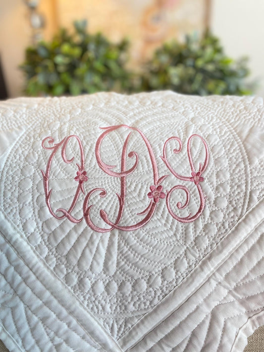 Close-up of a white quilt featuring an elegant pink embroidered monogram with floral accents, showcasing the intricate stitching on the Personalized Vintage Satin Monogram Baby Quilt | Custom Embroidered Heirloom Blanket.