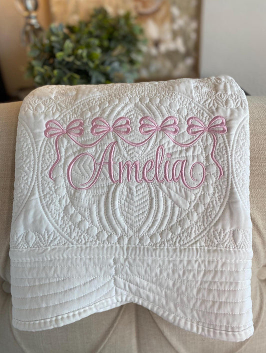 White quilted baby blanket with intricate stitching and delicate pink embroidered bows above a custom name, showcasing the Personalized Pink Bow Baby Quilt | Custom Name Heirloom Blanket for Girls.