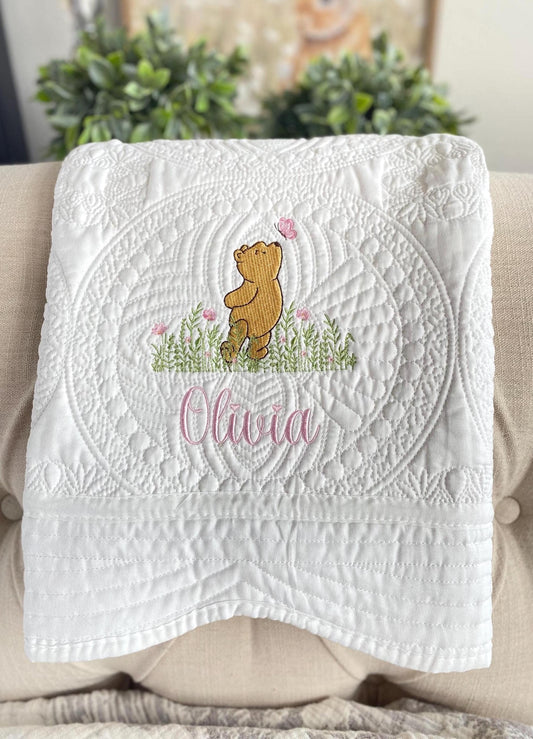 Personalized Classic Pooh Bear Meadow & Butterfly Baby Quilt | Custom Name Heirloom Blanket featuring detailed embroidery of Pooh Bear, flowers, butterfly, and the name Olivia on a white quilted background.