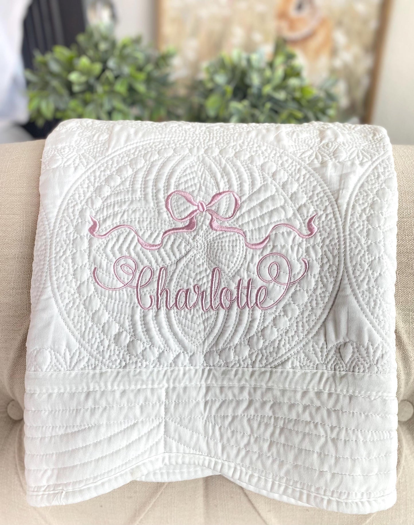 White quilt featuring intricate stitching and an embroidered pink ribbon bow with the name Charlotte, showcasing the Personalized Pastel Pink Ribbon Bow Baby Quilt | Custom Name Heirloom Blanket for Girls design.