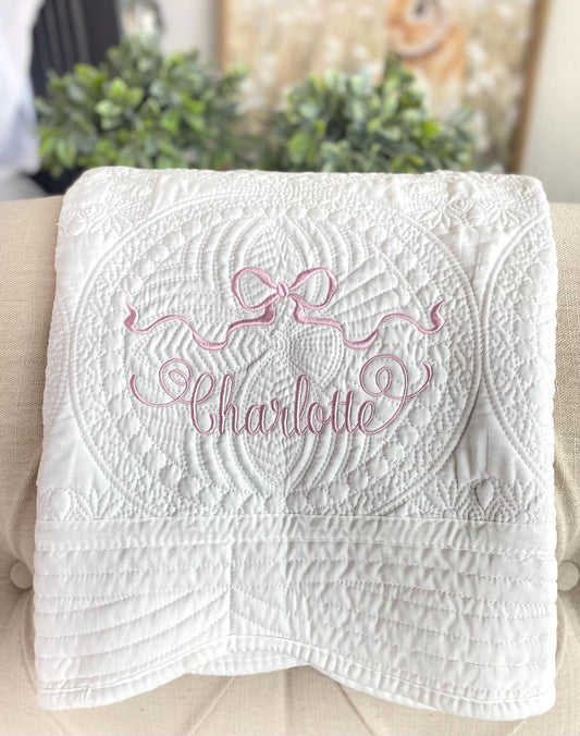White quilt featuring intricate stitching and an embroidered pink ribbon bow with the name Charlotte, showcasing the Personalized Pastel Pink Ribbon Bow Baby Quilt | Custom Name Heirloom Blanket for Girls design.