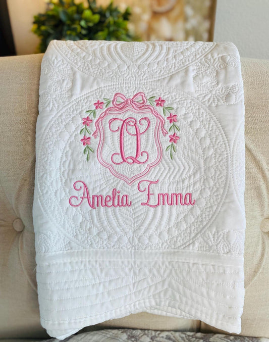 White quilt with intricate stitching, featuring a pink monogram inside a floral wreath with a bow, and the name Amelia Emma embroidered beneath, Personalized Floral Wreath & Bow Monogram Baby Quilt | Custom Name Heirloom Keepsake.