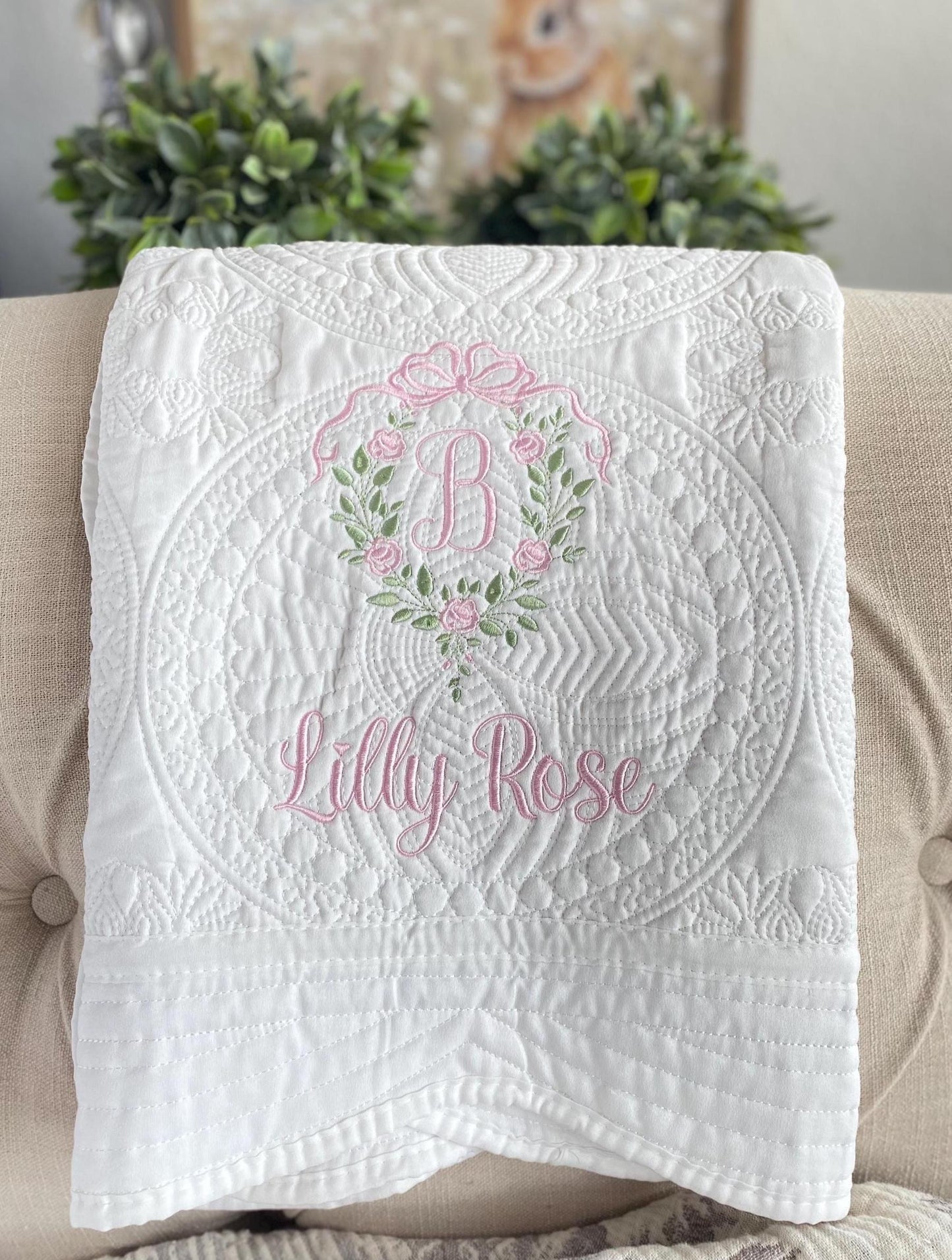 Personalized Rose Vine Bow Crest Monogram Baby Quilt | Custom Embroidered Heirloom Blanket featuring delicate pink embroidery of a floral vine wreath with a bow and the name Lilly Rose on a white quilted background.