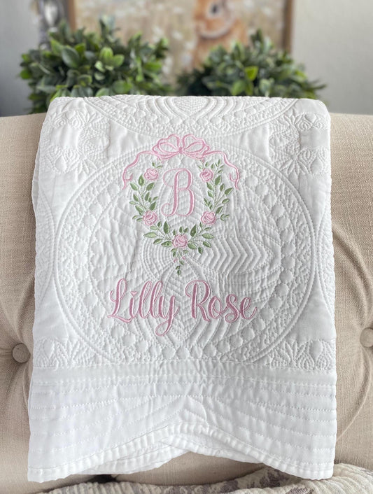 Personalized Rose Vine Bow Crest Monogram Baby Quilt | Custom Embroidered Heirloom Blanket featuring delicate pink embroidery of a floral vine wreath with a bow and the name Lilly Rose on a white quilted background.