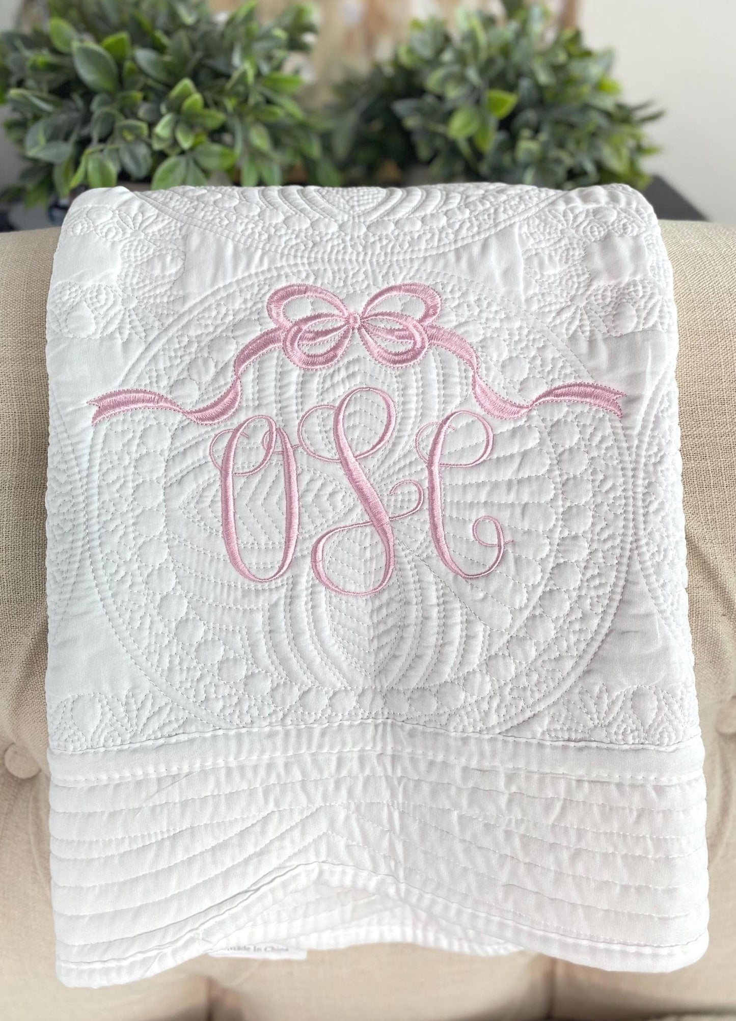 Soft white quilt with intricate stitching and a pink embroidered bow above a monogram, showcasing the Personalized Bow Monogram Baby Quilt | Custom Name Pink Heirloom Blanket perfect for a newborn gift.