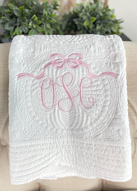 Soft white quilt with intricate stitching and a pink embroidered bow above a monogram, showcasing the Personalized Bow Monogram Baby Quilt | Custom Name Pink Heirloom Blanket perfect for a newborn gift.
