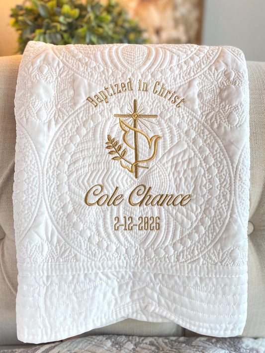 White quilt with intricate quilted patterns featuring an embroidered gold dove, cross, olive branch, and text "Baptized in Christ" with custom name "Cole Chance" and date "2-12-2026" on the Personalized Baptized in Christ Dove & Cross Baby Quilt | Custom Name & Date Christening Heirloom Blanket.