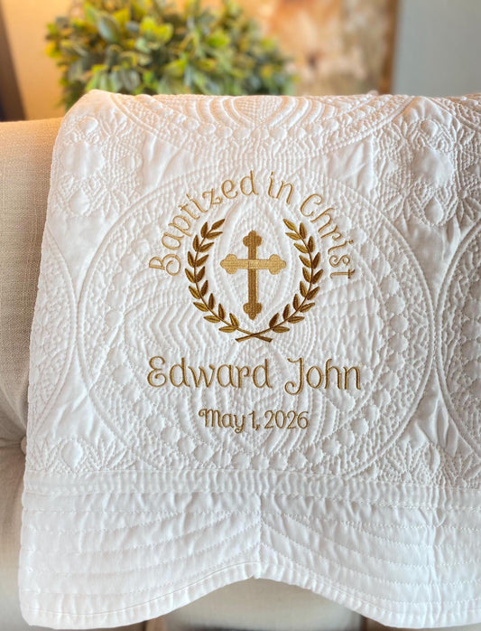 White quilt with intricate stitching featuring a gold embroidered cross and laurel wreath, personalized with name and date, titled Personalized Baptized in Christ Laurel Cross Baby Quilt | Custom Embroidered Christening Heirloom Blanket