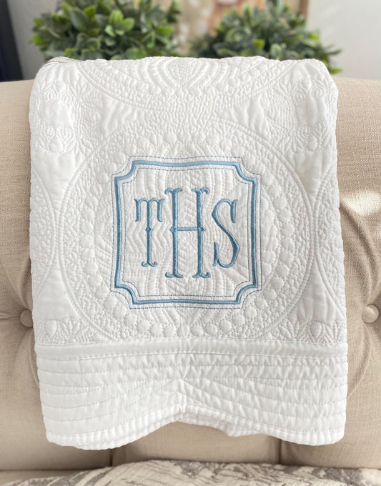 White quilt featuring a blue embroidered monogram with intricate stitching displayed on a beige tufted chair, showcasing the Personalized Square Frame Monogram Baby Quilt | Classic Embroidered Heirloom Keepsake.