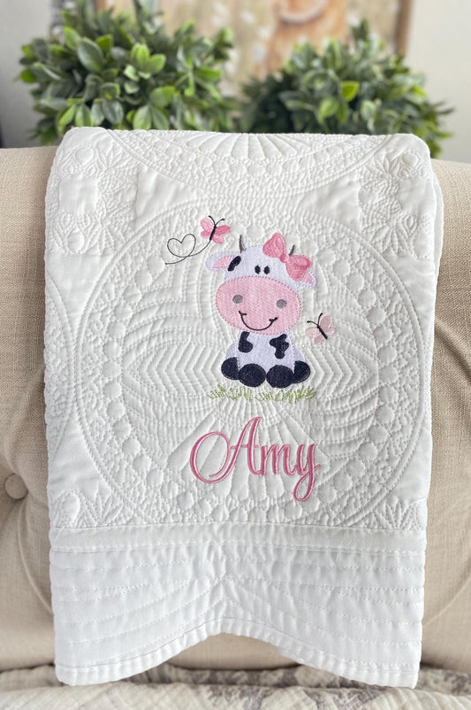 White quilt featuring a cute embroidered cow with pink bows, butterflies, and the name Amy in pink script, showcasing the Personalized Embroidered Cow Baby Quilt | Custom Name Blanket | Heirloom Keepsake.