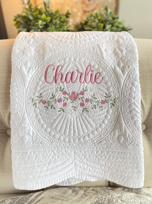 White quilt with intricate floral and scallop embroidery, personalized with the name Charlie in pink script, showcasing the Personalized Delicate Floral Rose Swag Baby Quilt | Custom Embroidered Heirloom Blanket.