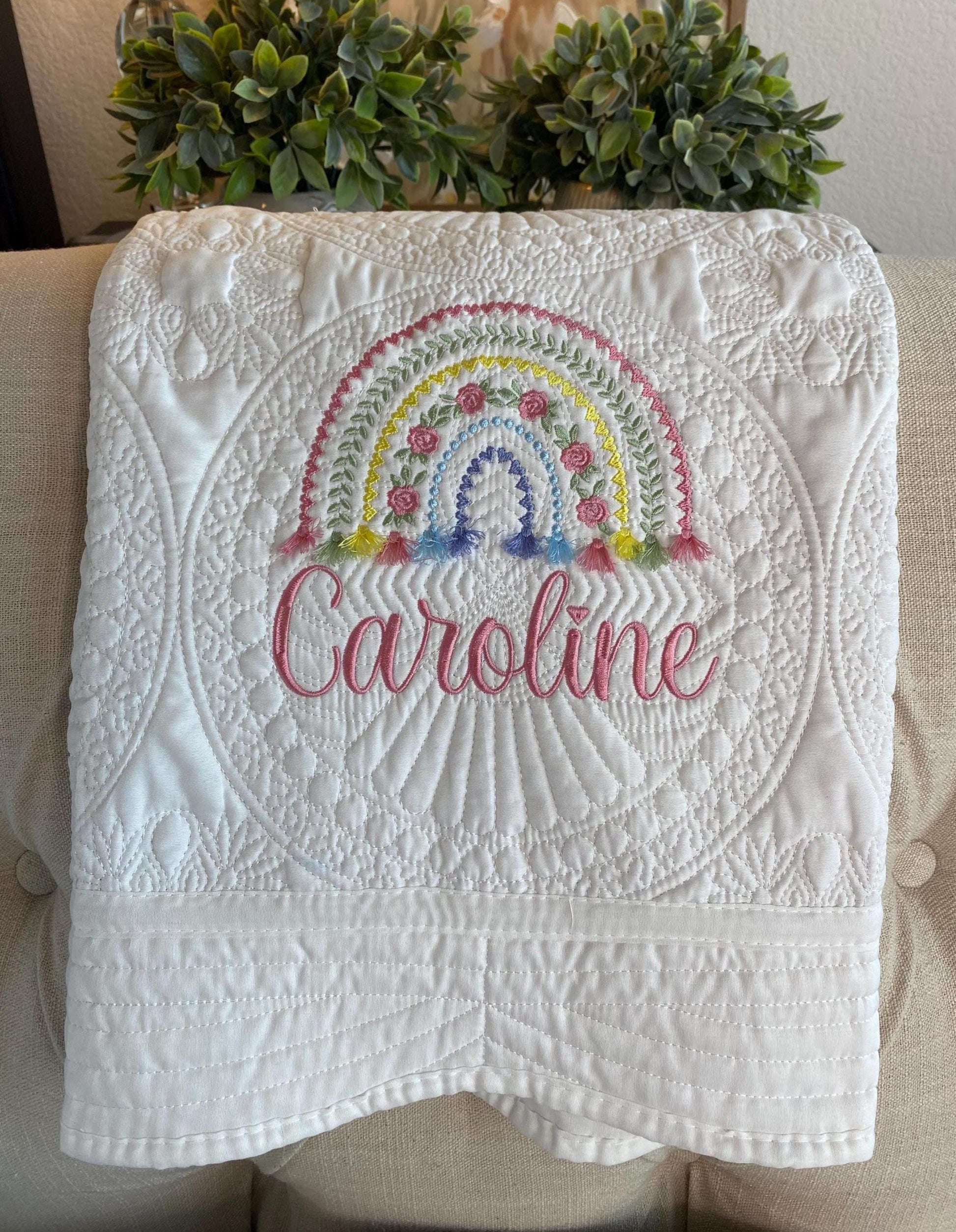 Personalized Boho Rainbow Baby Quilt | Custom Name Heirloom Blanket for Girls featuring a white quilt with intricate stitching, embroidered rainbow, and the name Caroline in pink cursive font.