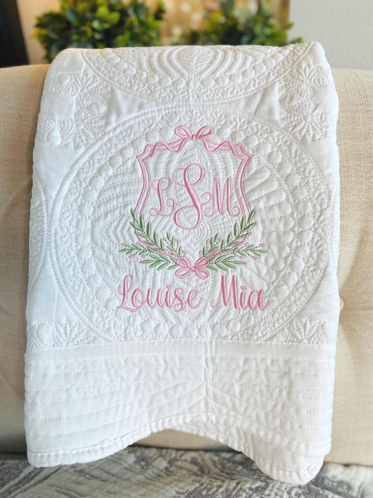 Personalized Olive Branch & Bow Crest Monogram Baby Quilt | Custom Embroidered Heirloom Blanket with delicate pink embroidery, featuring a monogram crest, olive branch, bow, and the name Louise Mia on a white quilted fabric.