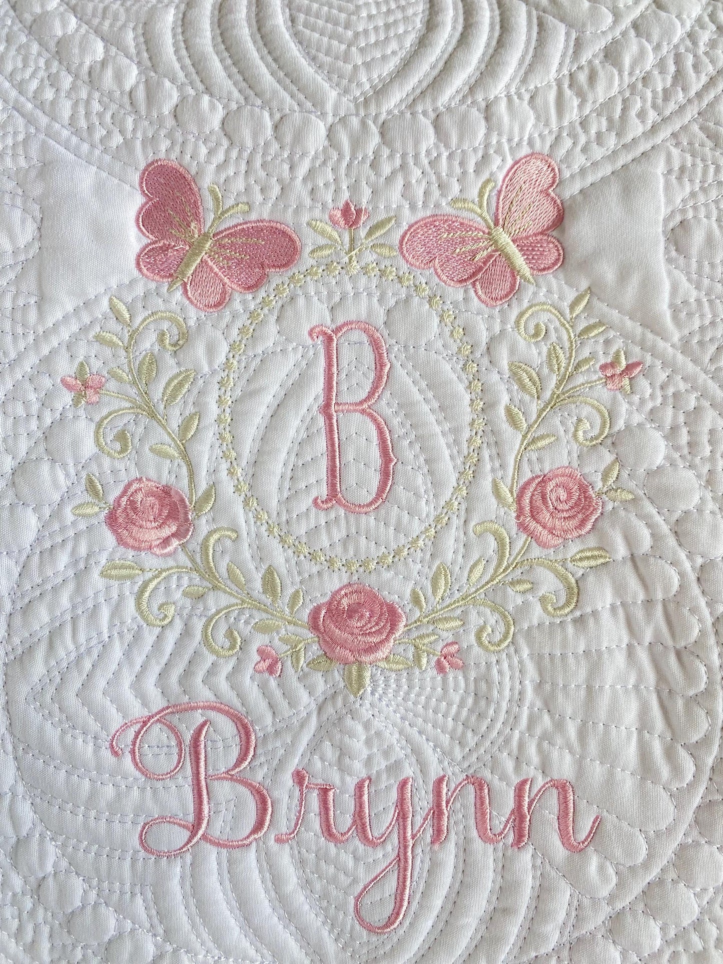 Close-up of a white quilt with intricate stitching featuring pink embroidered butterflies, roses, a monogram "B," and the name Brynn in elegant script on the Personalized Embroidered Rose & Butterfly Monogram Baby Quilt | Heirloom Keepsake