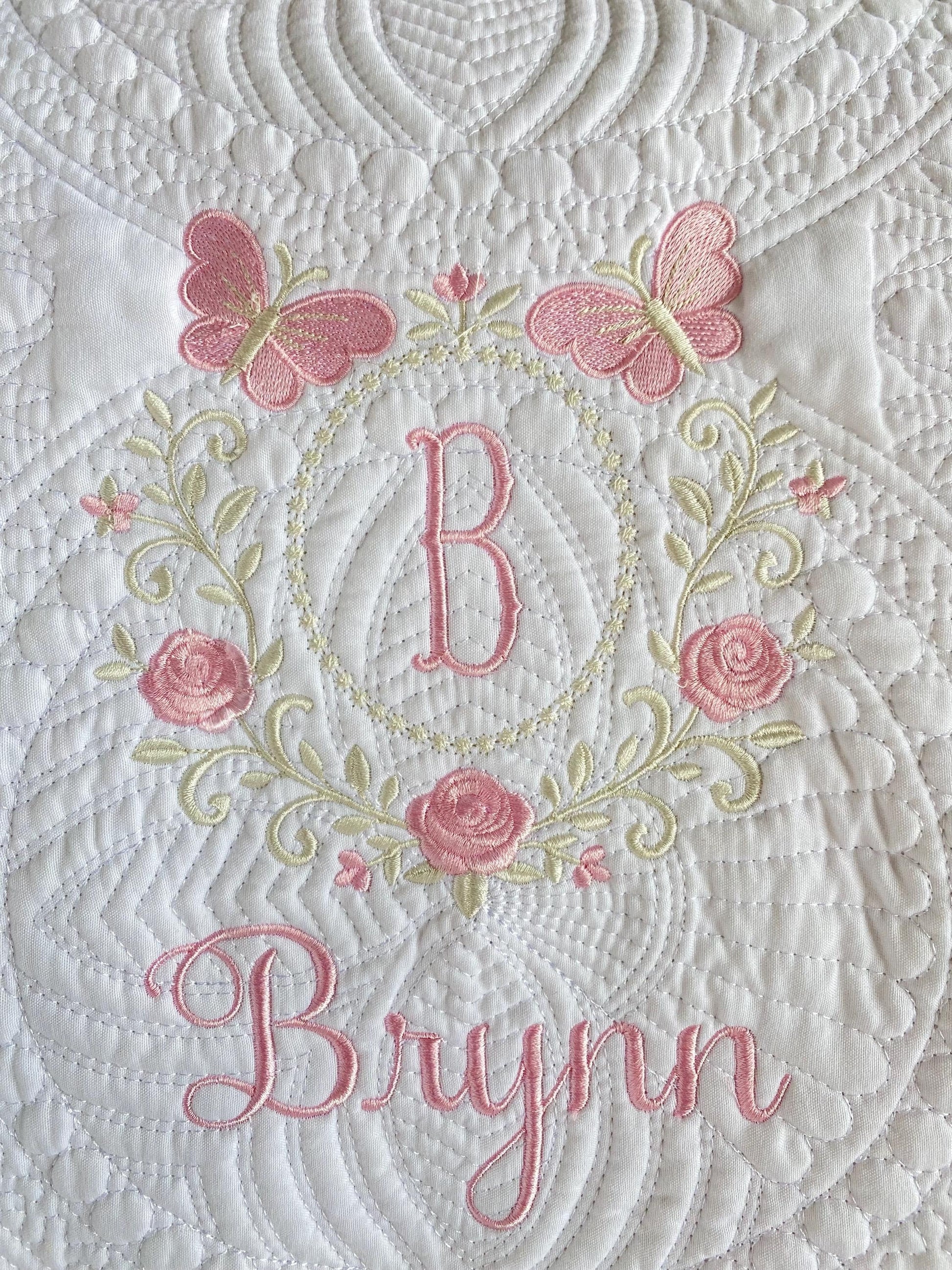 Close-up of a white quilt with intricate stitching featuring pink embroidered butterflies, roses, a monogram "B," and the name Brynn in elegant script on the Personalized Embroidered Rose & Butterfly Monogram Baby Quilt | Heirloom Keepsake