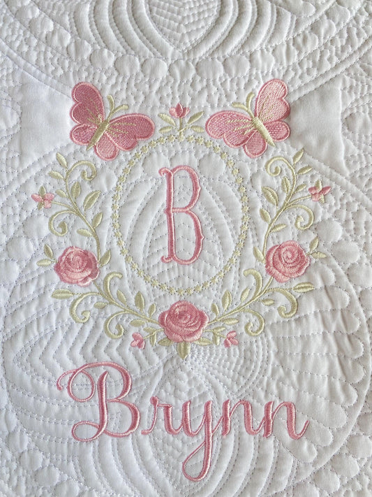Close-up of a white quilt with intricate stitching featuring pink embroidered butterflies, roses, a monogram "B," and the name Brynn in elegant script on the Personalized Embroidered Rose & Butterfly Monogram Baby Quilt | Heirloom Keepsake